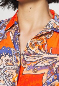 Kortärmad skjorta i livlig orange med intrikata paisleymönster i blått och vitt, med spetsig krage och knapprader.