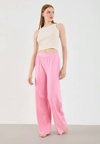 Jonge vrouw die blootsvoets staat in een mouwloze witte crop top en losse roze wijde broek tegen een effen achtergrond.