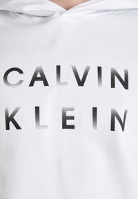 Bela kapuca z drsno tkanino in bold črnim gradientnim besedilom "CALVIN KLEIN", z detajli vidnega šivanja in z vrvico za nastavitev kapuce.