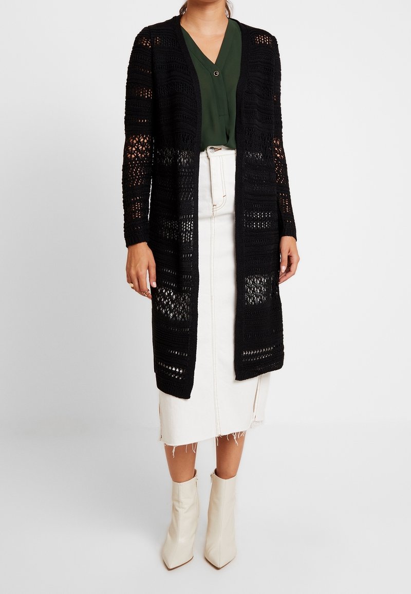 Black open-knit cardigan with long sleeves, featuring a textured pattern; paired with a white denim skirt and cream ankle boots.
