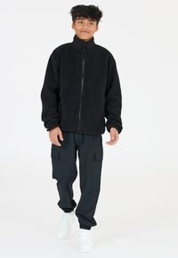 Veste en polaire noire avec un col haut, fermeture éclair sur le devant et poches latérales, associée à un pantalon cargo foncé et des baskets blanches.