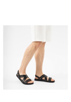 Gambe con pantaloncini bianchi e sandali di pelle neri con cinturini incrociati e fibbia, in piedi su sfondo bianco.