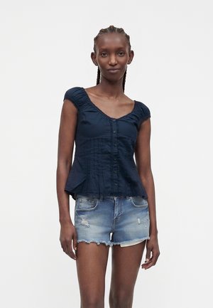 Donna con i capelli intrecciati che indossa una camicetta blu navy a maniche corte con bottoni e shorts di jeans usurati, in piedi davanti a uno sfondo semplice.