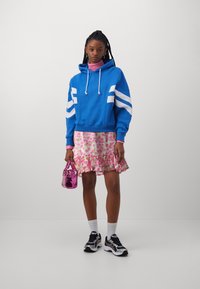 Sudadera con capucha azul y rayas blancas, falda rosa floral con volantes, bolso rosa brillante, zapatillas negras y blancas, calcetas blancas de canalé.