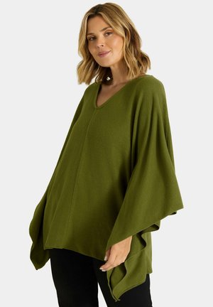 Vrouw met schouderlang blond haar die een losse olijfgroene poncho draagt over een zwarte broek, staand tegen een effen achtergrond.
