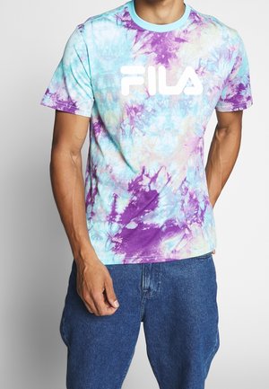 Mężczyzna ubrany w koszulkę FILA w kolorze niebieskim, fioletowym i białym w stylu tie-dye oraz niebieskie dżinsy, stojący na tle jednolitego jasnego tła.