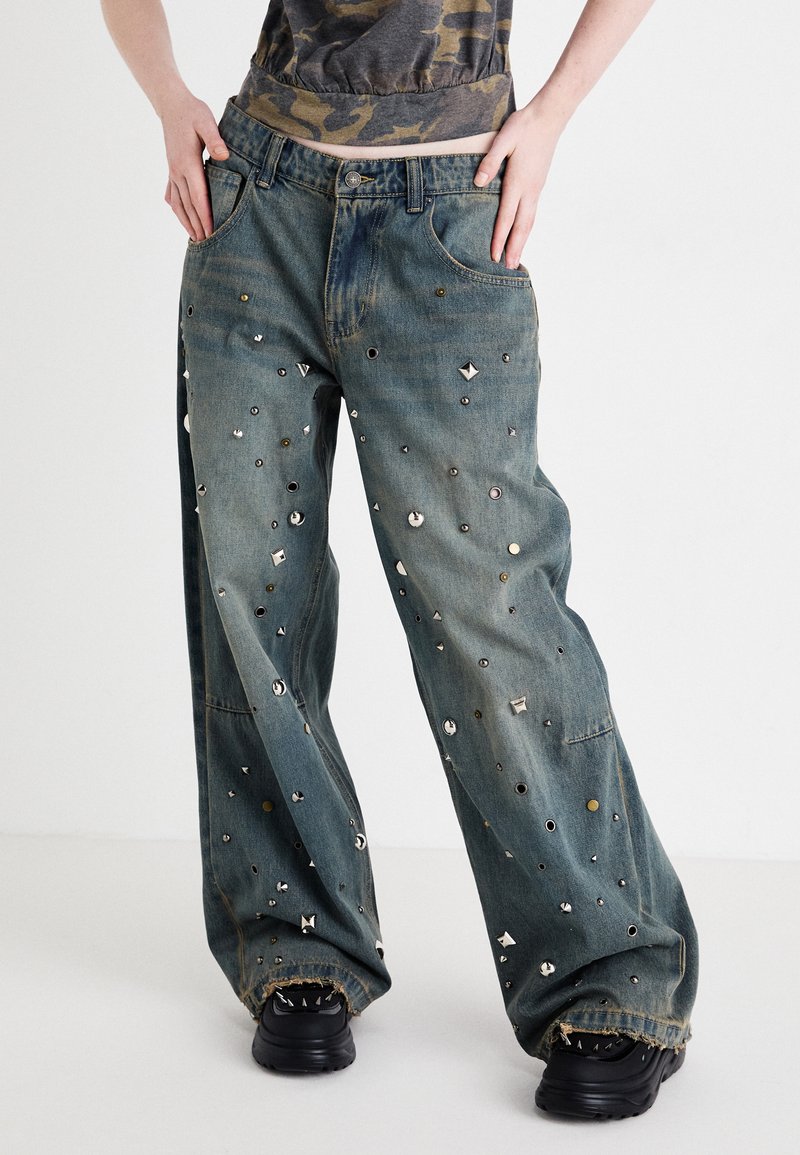 Calças de ganga largas de tecido azul, com vários tachas metálicas e buracos, apresentando uma bainha ligeiramente desfiada e dois bolsos frontais. Calçado preto visível.