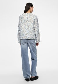 Blouse à manches longues florale avec un fond bleu clair, ornée de fleurs jaunes et bleues, et de poignets volantés. Associée à un jean large bleu clair.
