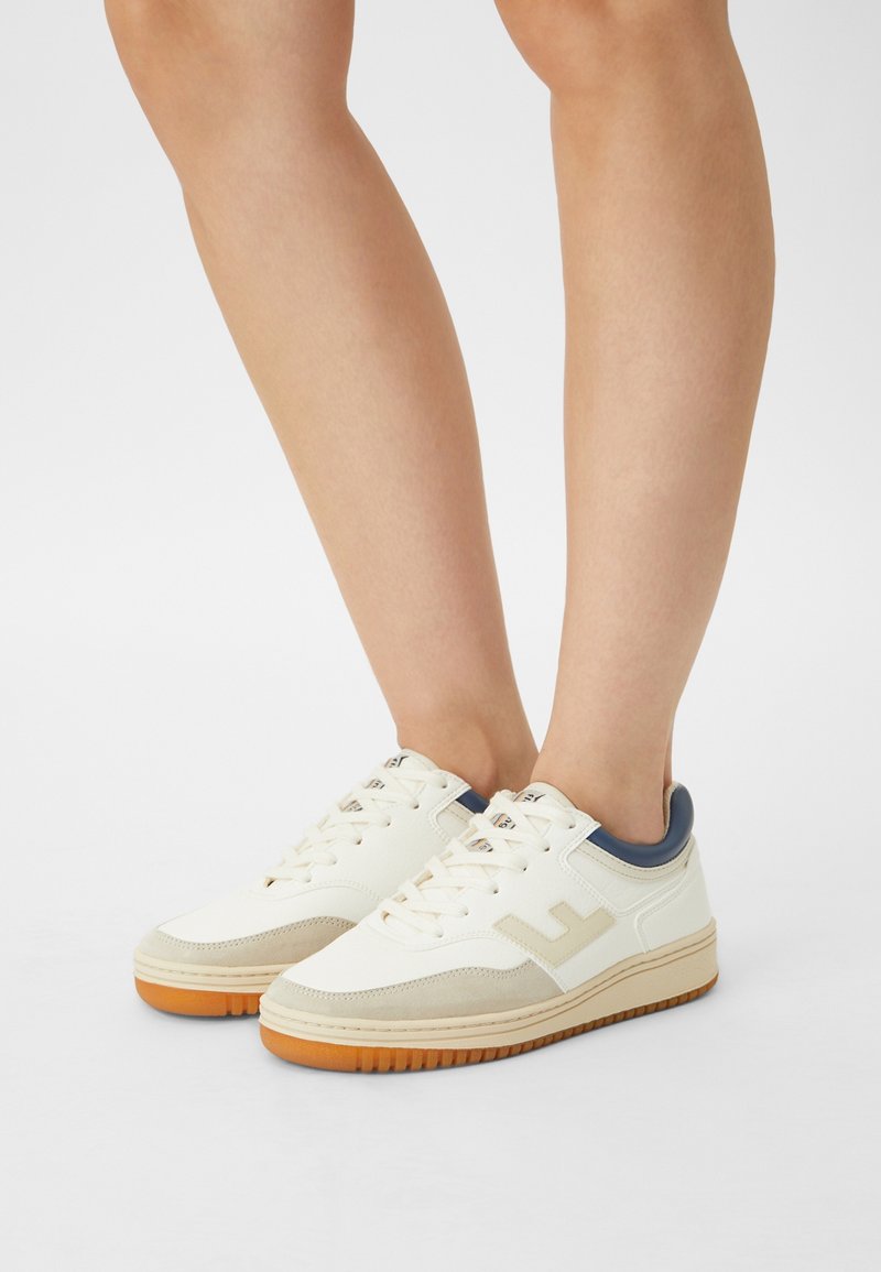 Sneakers bianchi con tomaia in pelle liscia, dettagli in suede beige chiaro e tallone blu navy. Suola in gomma con grip testurizzato.