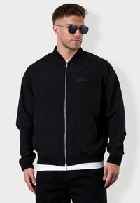 Schwarze Bomberjacke aus leichtem Stoff, mit einem Frontreißverschluss, gerippten Bündchen und einem rechteckigen Logo-Patch auf der Brust.