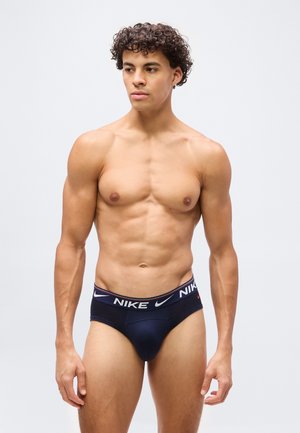 Svalnatý muž s kučeravými vlasmi v tmavých slipoch značky Nike, stojaci na svetlosivom pozadí, pozerajúci sa stranou.