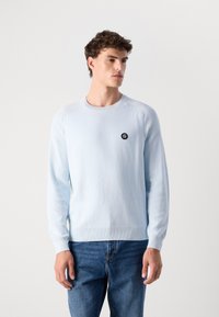 Light blue knitted sweater with a crew neck and ribbed cuffs. Features a small black logo on the chest. Paired with blue jeans.