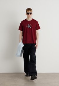 Bordo pamučna majica s grafičkim logotipom, crne hlače širokih nogavica, crne cipele i svijetloplava dukserica koja je prebačena preko ruke.