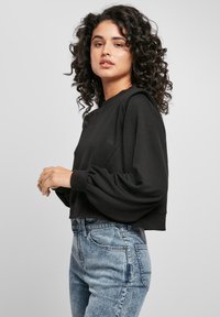 Femme aux cheveux bouclés portant un pull noir à manches longues bouffantes et un jean taille haute bleu clair, se tenant contre un fond uni.