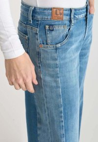 Close-up van lichtblauwe spijkerbroek met zichtbare voorzak, riemlussen, logo-patch, en een hand die naast de heup rust.