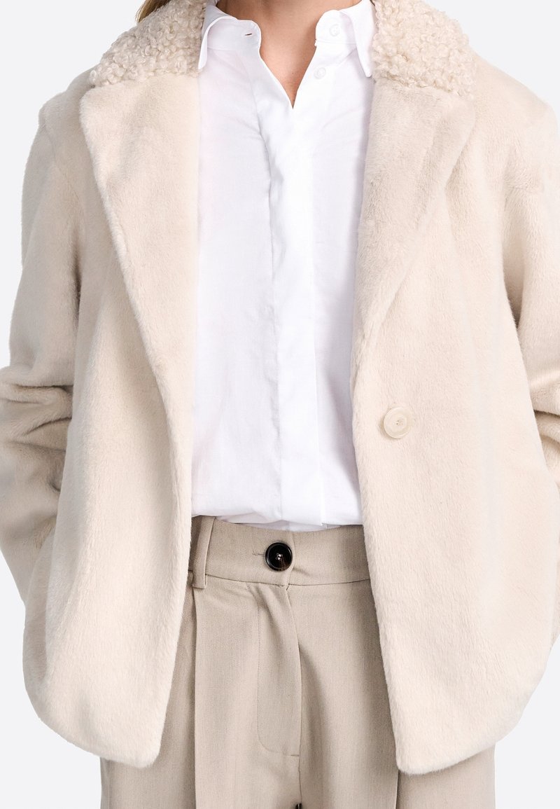 Femme portant un manteau en fausse fourrure crème avec un col texturé, une chemise blanche à boutons et un pantalon beige taille haute avec un bouton noir.