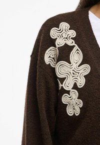 Tumši brūns džemperis ar baltām abstraktām ziedu izšuvumiem uz pleca, uzvilkts virs baltas krekla.