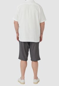 Man in een wit overhemd met korte mouwen, grijze broek tot de knie en witte sneakers, staand met zijn rug naar de camera tegen een effen achtergrond.