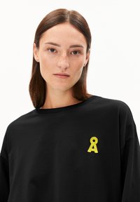 Schwarzes T-Shirt mit kurzen Ärmeln aus weicher Baumwolle, versehen mit einem gelben gestickten Design im linken Brustbereich. Einfacher und lässiger Schnitt.