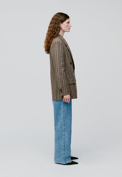 Blazer de cuadros marrón con un corte entallado, combinado con jeans de mezclilla claros de tiro alto y zapatos negros de punta. Tela texturada y diseño estructurado.