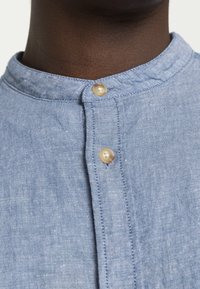 Ljusblå linneskjorta med rundad krage, med två ljusa knappar vid halsen och en strukturerad tyg-yta.