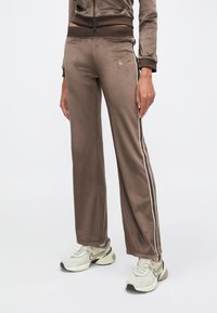 Άτομο που φοράει καφέ βελουτέ παντελόνι φόρμας με λευκές ρίγες στο πλάι και μπεζ αθλητικά παπούτσια Nike, στέκεται μπροστά σε απλό φόντο.