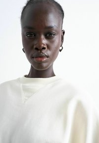 Femme noire aux cheveux naturels, portant des boucles d'oreilles créoles et un sweat-shirt blanc, regardant directement l'appareil photo sur un fond uni.