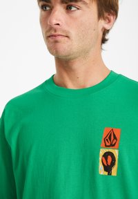 T-shirt en coton vert avec un graphique sur la poitrine gauche, sur un fond orange et jaune, affichant des formes abstraites en noir.