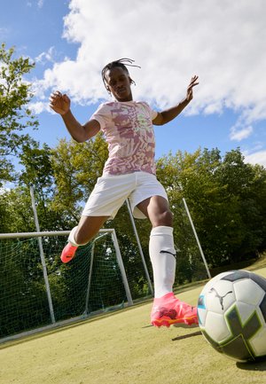 Jucător îmbrăcat într-o cămașă roz deschis cu un model și pantaloni scurți albi lovește o minge de fotbal. Poartă ghete roz și ciorapi albi până la genunchi. Pe fundal se văd un teren verde și un stâlp de poartă.