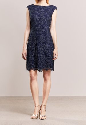 Kobieta w granatowej koronkowej sukience bez rękawów i złotych sandałach na wysokim obcasie, stojąca na tle jasnego, jednolitego tła.