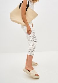 Beige vævet tote taske med sorte håndtag, hvid perforeret strikkjole og beige quilted slip-on sandaler med tykke såler.