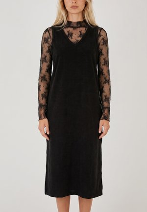 Donna che indossa un vestito nero senza maniche sopra una maglia in pizzo trasparente a maniche lunghe con motivi floreali, in piedi su uno sfondo semplice.