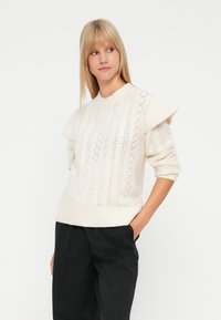 Blondynka w białym swetrze z teksturowanego dzianiny z bufkami na ramionach i czarnych spodniach w paski, stojąca z jedną ręką w kieszeni.