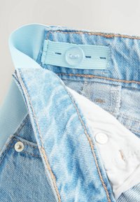 Jeans de mezclilla azul claro con una cinturilla suave, cierre de botón y detalle de costura dorada a lo largo de las costuras. El interior revela un forro blanco.