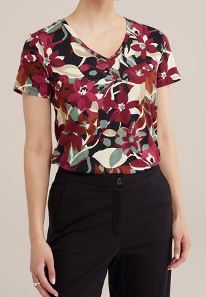Woman wearing a short-sleeve V-neck floral top with red, green, and cream flowers paired with black high-waisted pants.