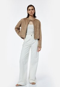 Beige Velourjacke mit rundem Halsausschnitt, Reißverschluss und langen Ärmeln, kombiniert mit weißen weit geschnittenen Jeans und hellen Sandalen.