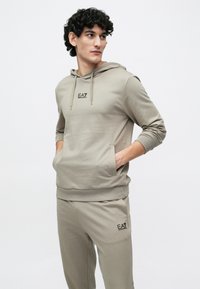 Férfi bézs színű EA7 Emporio Armani kapucnis pulóverben és hozzáillő melegítőnadrágban, kezeit a zsebében tartva, oldalra néz egy egyszínű háttér előtt.