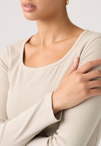 Μπεζ μακρυμάνικη μπλούζα με ριμπ ύφασμα και στρογγυλή λαιμόκοψη, με εφαρμοστά μανίκια και μαλακή υφή, εμφανιζόμενη στο πάνω μέρος του σώματος ενός μοντέλου.