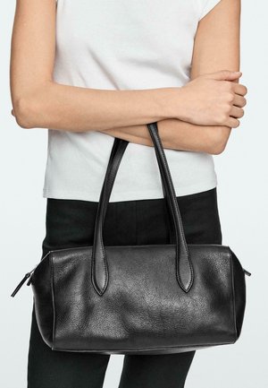 Personne portant un haut blanc et un pantalon noir tenant un sac à main en cuir noir avec deux poignées devant sa taille.