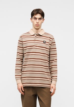 Jauns vīrietis, tērpts bēšā un brūnā svītrainā garroku polo krekliņā un brūnās biksēs, stāvot pret vienkāršu gaišu fonu.