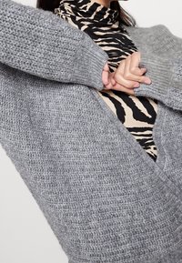 Grå stickad cardigan med texturmönster, lösa ärmar och V-ringning, lager på en topp med svart och beige zebramönster.