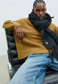 Strikket sennepsfarvet sweater og sort uld tørklæde, sidder på en sort læderstol. Blå denim jeans med lys vask og let slid.