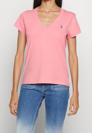 Różowy bawełniany t-shirt z dekoltem w serek, krótki rękaw, luźny krój i małe niebieskie logo na lewej piersi. Noszony z dżinsami.