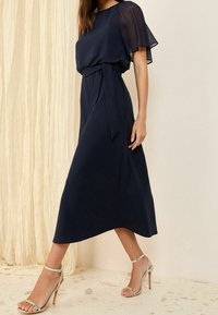 Navy blue midi dress with a sheer short-sleeve overlay, belted waist, and smooth fabric; paired with silver strappy high-heeled sandals.