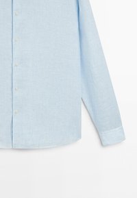 Chemise bleu clair à manches longues avec fines rayures verticales blanches, montrée du milieu du torse jusqu’au poignet sur fond blanc.