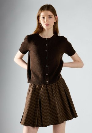 Giovane donna con lunghi capelli castani che indossa un maglione marrone scuro a maniche corte con bottoni e una gonna plissettata a quadri marrone.