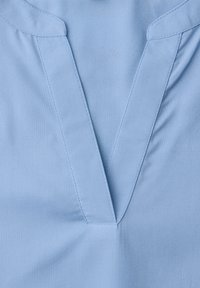 Vêtement bleu clair avec un col en V et des coutures visibles le long du col et de l'ouverture frontale.