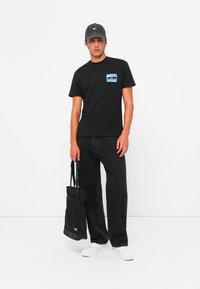 Schwarzes Baumwoll-T-Shirt mit Grafik auf der vorderen Tasche, kombiniert mit schwarzen weiten Jeans, weißen Sneakers und einer schwarzen Mütze, während er eine schwarze Tragetasche hält.