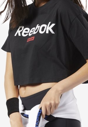 Kvinde iført en sort cropped Reebok t-shirt, sort håndledsbind og hvid jakke bundet om taljen mod hvid baggrund.