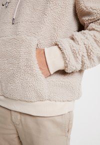 Beige Sherpa-Fleece-Hoodie mit Kängurutasche, elastischen Bündchen und einer weichen, strukturierten Oberfläche. Darunter trägt sie einen hellbeigen Pullover.